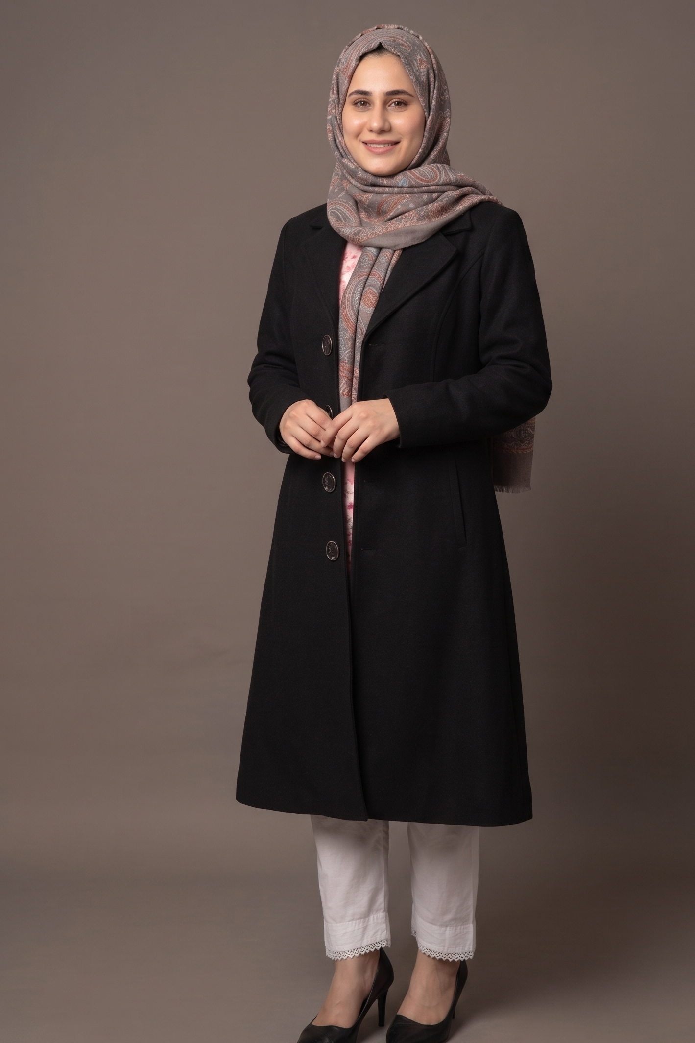 Woman wearing a black coat, white pants, and a patterned hijab on a gray background
